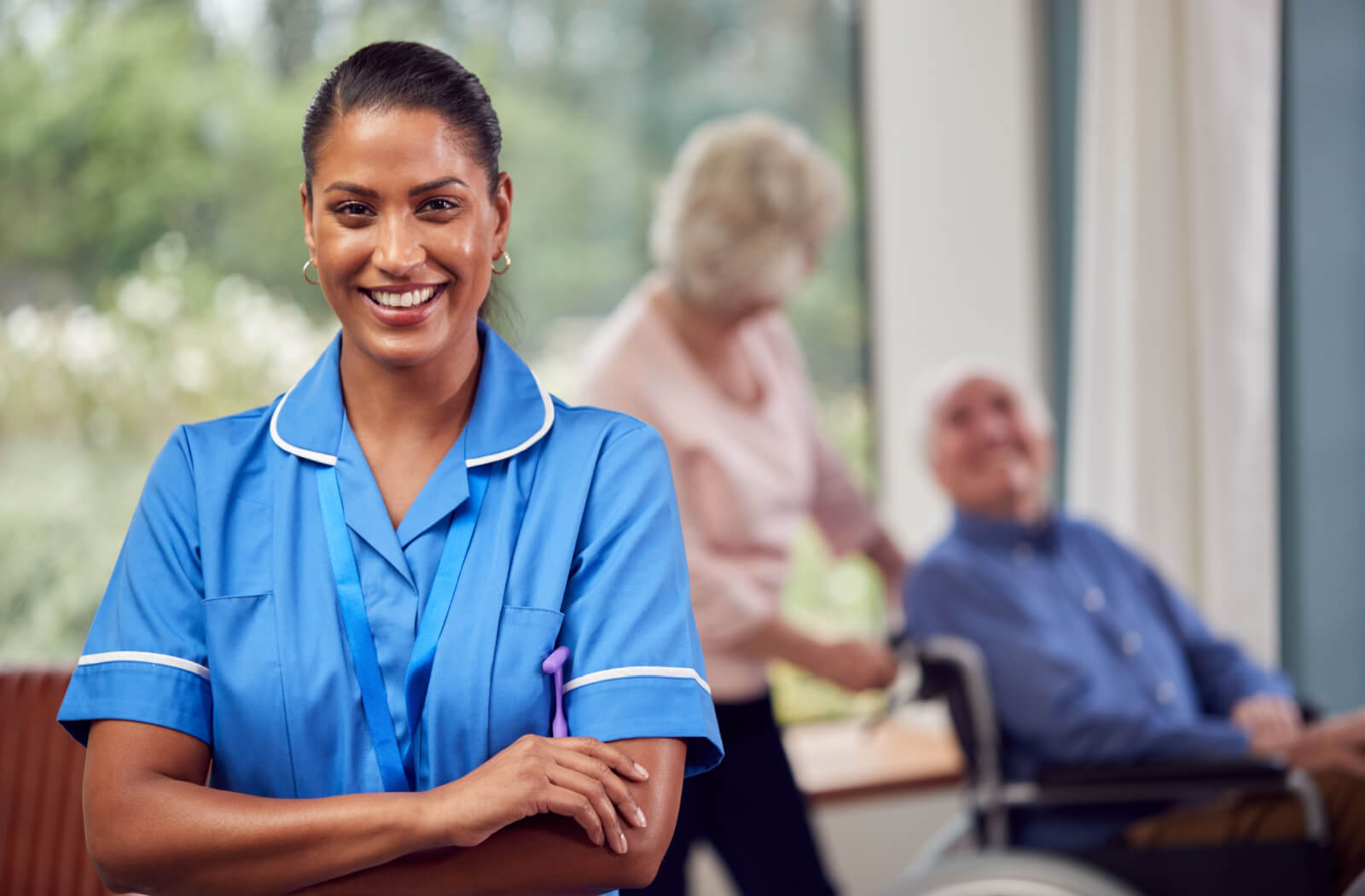 A staff member of a personal care home smiles in the foreground while two happy residents interact in the background.