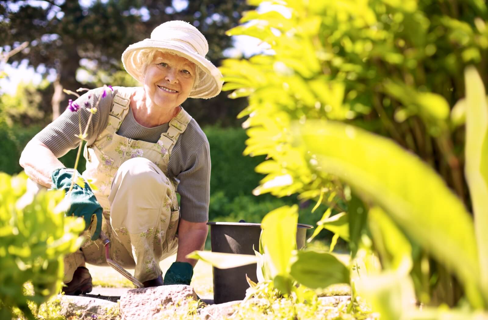 An older adult smiling while crouching in their garden and digging with an ergonomic trowel.