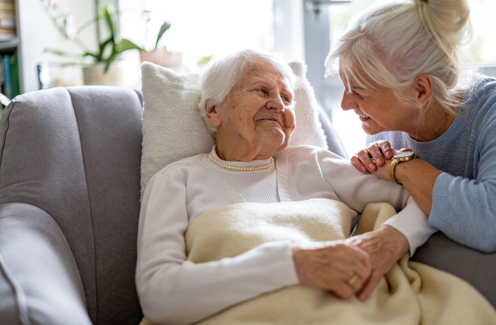 An older adult smiles while sitting on the couch during a visit with their adult child in memory care